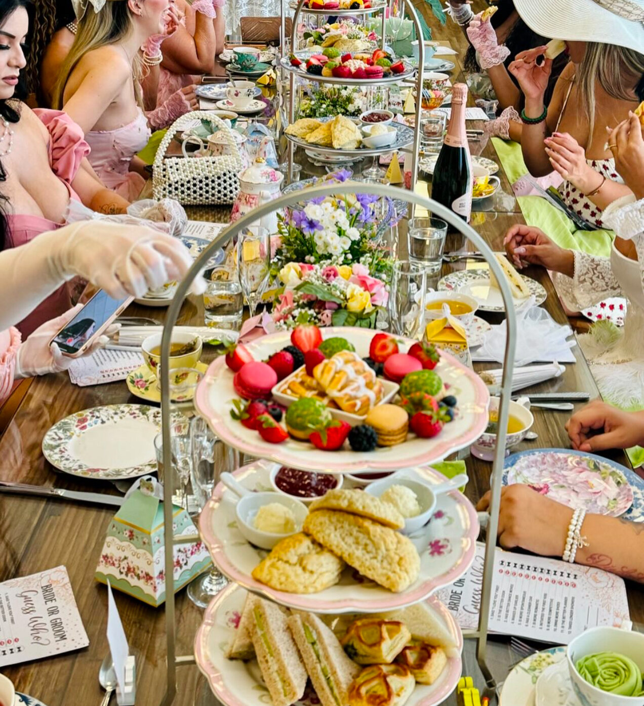 A lavish tea party table with women dressed in pastel and floral outfits. The table is set with tiered trays of pastries, cakes, scones, finger sandwiches, fruits, and tea cups, creating an elegant, festive atmosphere.
