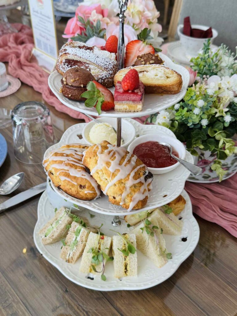 A three-tiered tray holds assorted pastries, scones with icing, jam, cream, and finger sandwiches. The display is set on a wooden table with flowers, glassware, and plates, creating an elegant afternoon tea setting.
