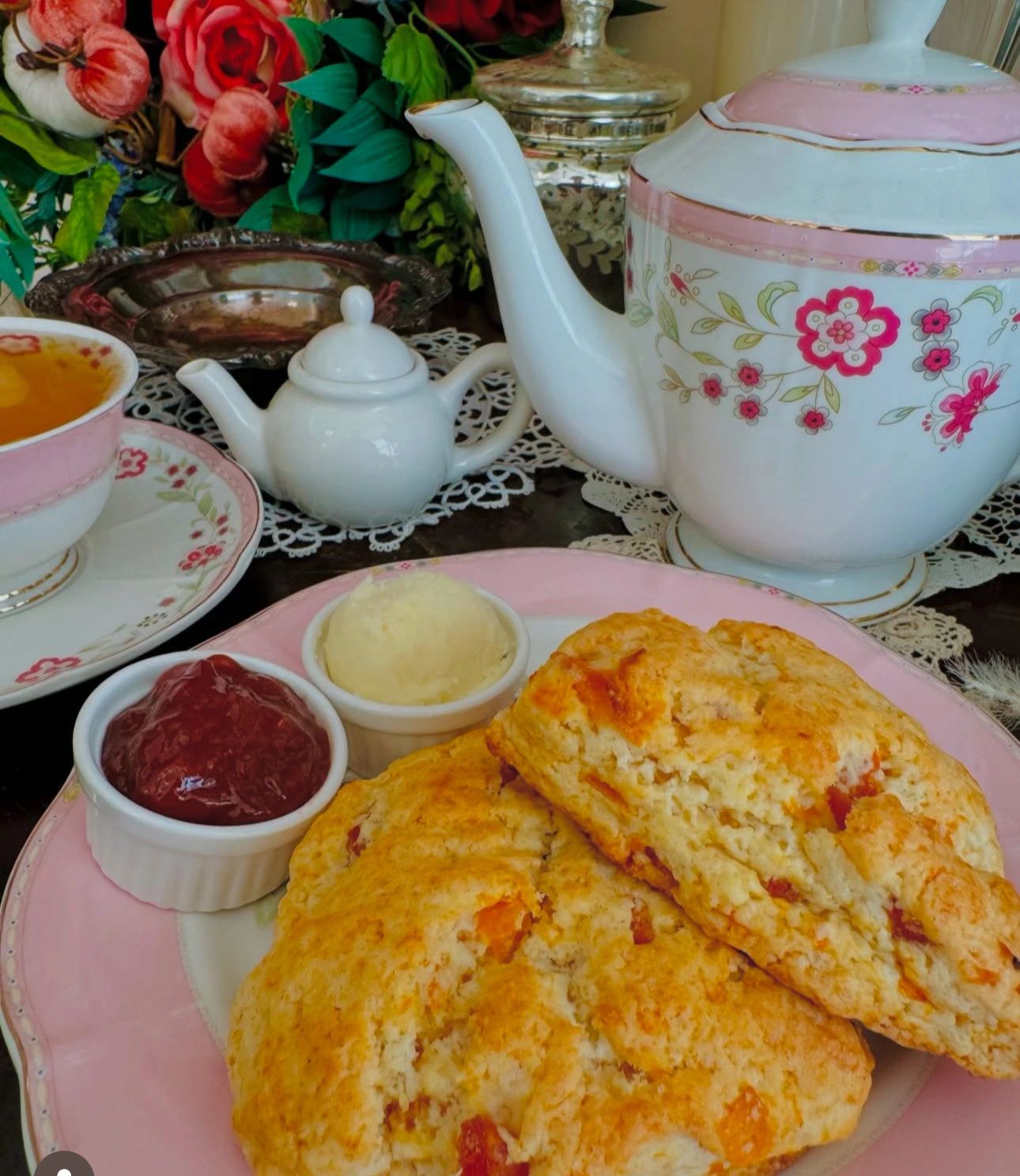 A plate with two fruit scones, clotted cream, and jam sits next to a pink teacup filled with tea and a white teapot with floral patterns, set on a lace tablecloth with flowers in the background.