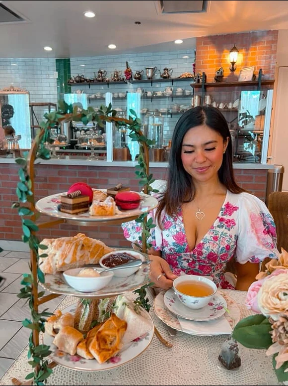 A woman in a floral dress sits at a table in a café, enjoying afternoon tea with assorted pastries and desserts on a tiered tray, holding a teacup and smiling. The café is cozy with vintage decor.