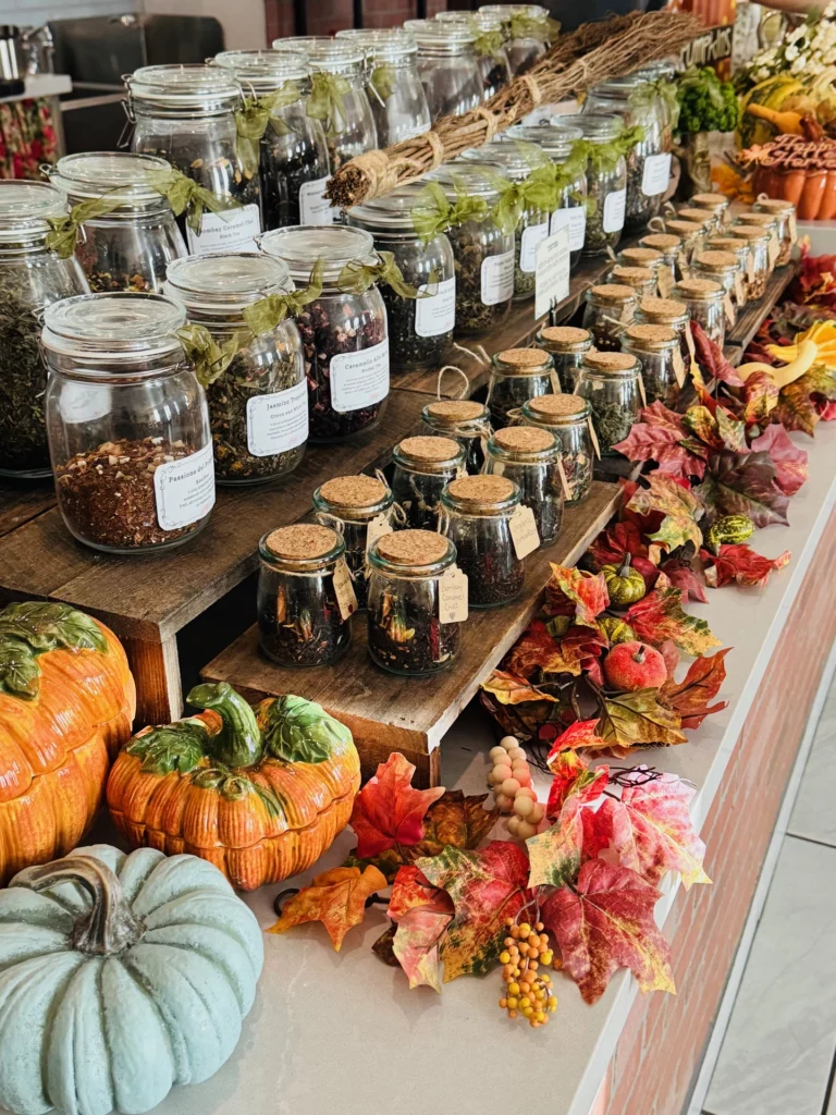 A display of glass jars filled with dried herbs and teas, arranged on wooden shelves. Autumn leaves, pumpkins, and faux fruit decorate the countertop, creating a festive fall atmosphere.