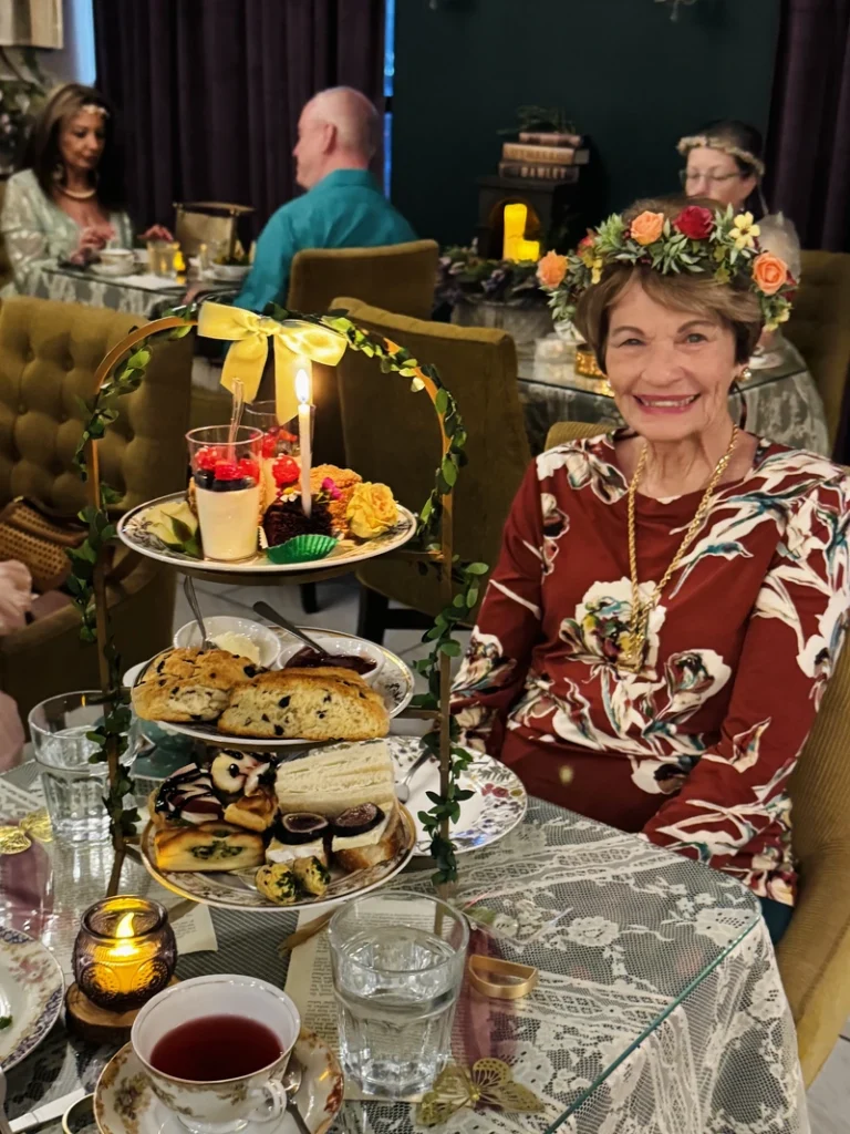 An older woman in a floral crown smiles at a tea table with a three-tiered tray of desserts and sandwiches. Others sit around her in a warmly lit, elegant room decorated with flowers and lace.