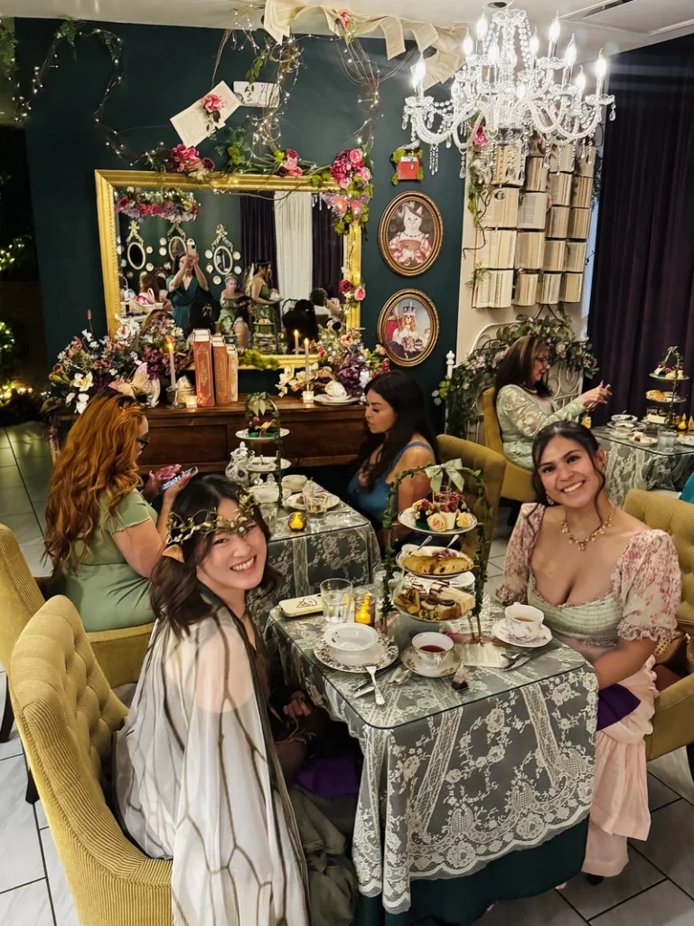 Four women in elegant dresses enjoy tea at a richly decorated, vintage-style tea room with floral arrangements, a chandelier, and a large ornate mirror reflecting the scene.