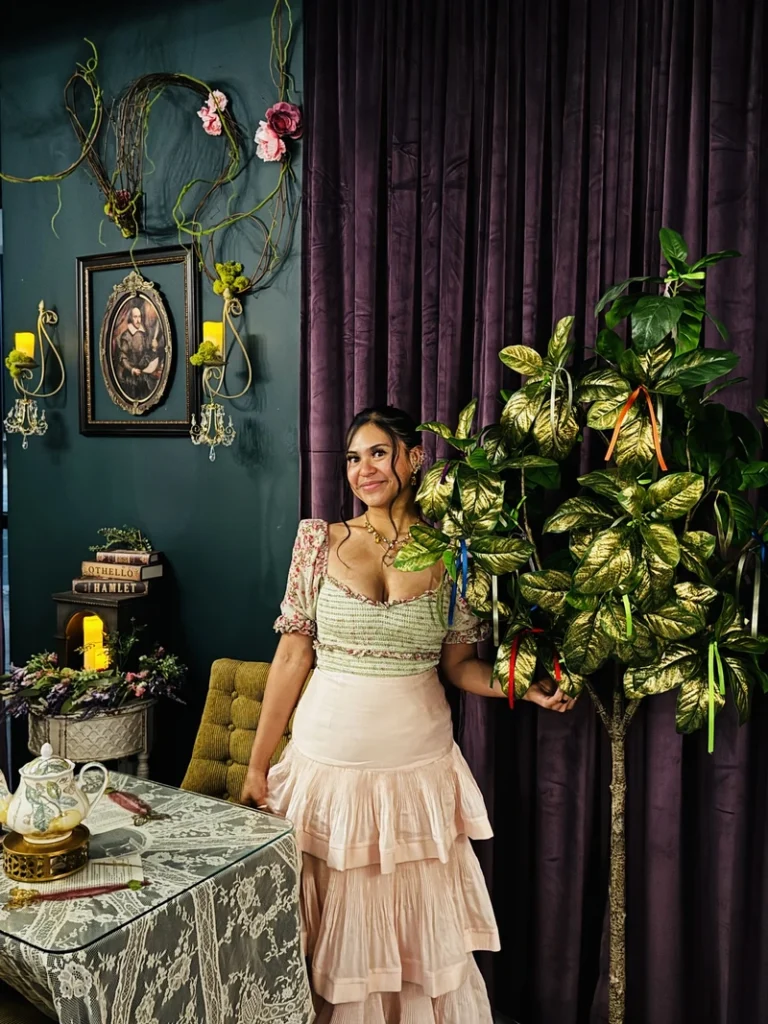 A woman in a floral top and tiered skirt stands beside a faux tree with gold leaves and ribbons, in a vintage-decorated room with a candlelit table, lace cloth, and framed art on a dark wall.